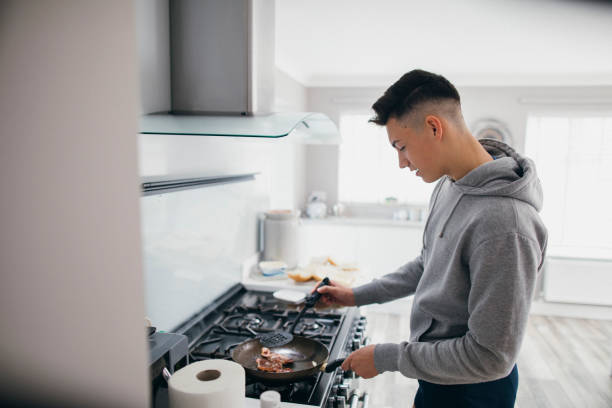 A teenage boy is frying bacon for a bacon sandwich.